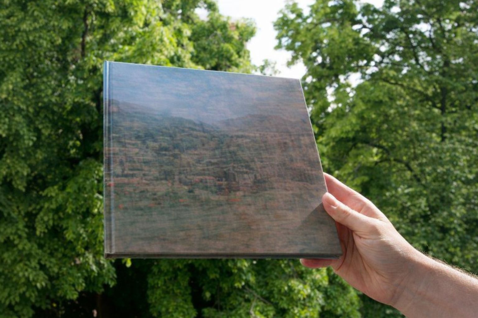 Eine Hand hält ein Buch mit einem Landschaftsbild vor grünem Laubwerk.